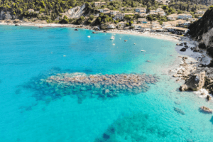 Sandstrand mit hellblauem Wasser neben einem Wohngebiet mit weißen Häusern mit Säulendächern und vielen grünen Bäumen und Pflanzen
