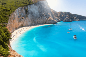 Sandstrand mit hellblauem Wasser neben einem hohen Felsen mit grünen Bäumen und Pflanzen