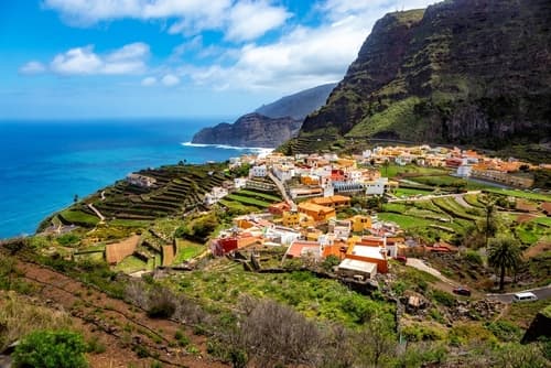 A picturesque village on La Gomera, Canary Islands, with colourful houses, terraced fields, and dramatic cliffs overlooking the deep blue ocean under a bright sky.
