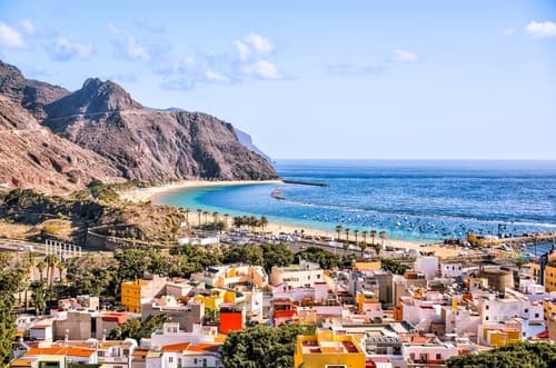 A vibrant coastal town on Tenerife, Canary Islands, featuring colourful buildings, a scenic beach, and rugged mountains set against the backdrop of a clear blue ocean.