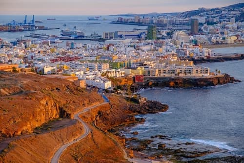 A coastal view of Las Palmas, Gran Canaria, Canary Islands, showcasing the bustling cityscape, harbour, and distant ships, set against rugged cliffs and the Atlantic Ocean.