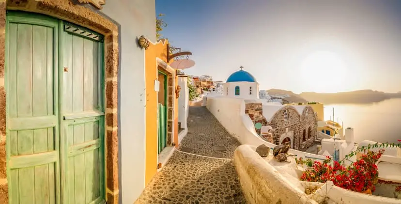 Vicolo di Oia a Santorini con chiesa a cupola blu, porte colorate e vista sulla caldera al tramonto