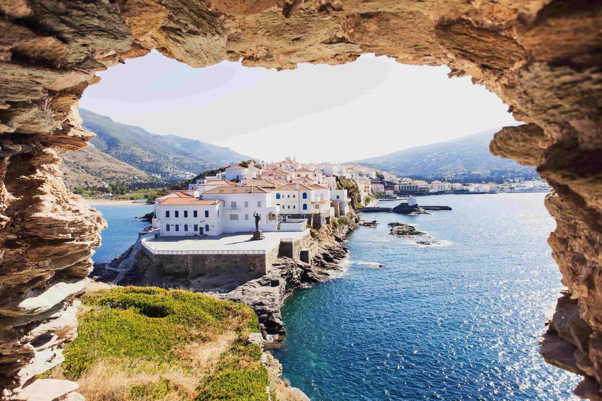 Άνδρος Χώρα – Πανοραμική θέα από φυσικό άνοιγμα στο βράχο προς το ακρωτήριο με τα λευκά αρχοντικά, την προτομή του Ναυτικού και τα καταγάλανα νερά του Αιγαίου, σε ένα από τα πιο γραφικά σημεία των Κυκλάδων.