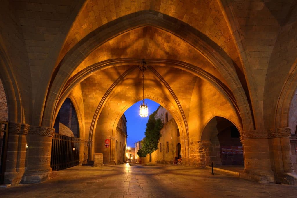 Η μεσαιωνική Ρόδος φωτισμένη στο σούρουπο: οι γοτθικές καμάρες δημιουργούν ένα σκηνικό γεμάτο μυστήριο, ενώ ο λιθόστρωτος δρόμος οδηγεί σε μια πόλη γεμάτη ιστορία.