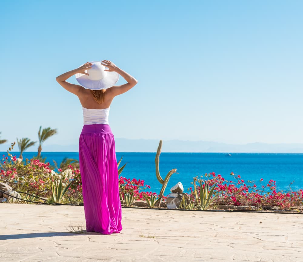 Giovane donna di spalle, vestita con una lunga gonna fucsia e un cappello bianco, mentre ammira il panorama sul mare turchese