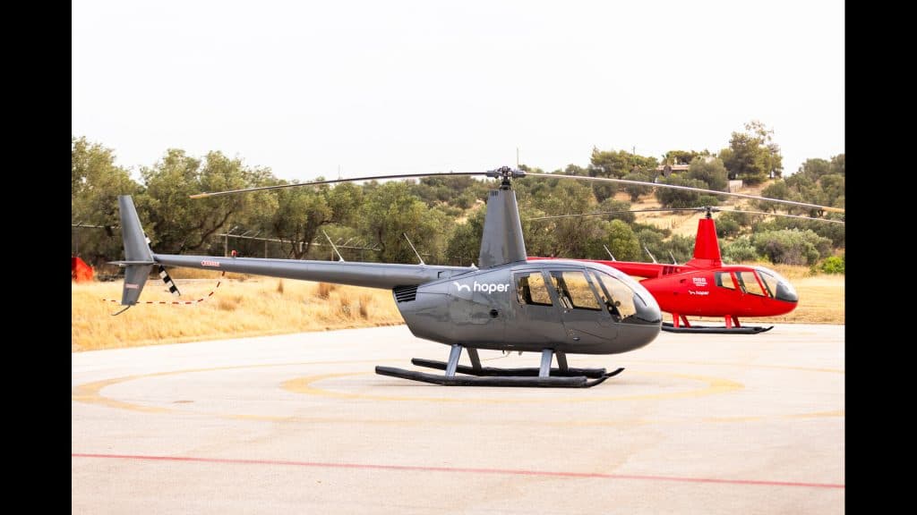 Two hoper helicopters—one gray and one red—parked on a helipad surrounded by dry grass and olive trees, ready for scheduled flights between the Greek islands.