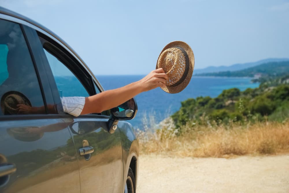 Persona che sporge il braccio fuori dal finestrino dell’auto sventolando un cappello, con vista panoramica sul mare e paesaggio mediterraneo