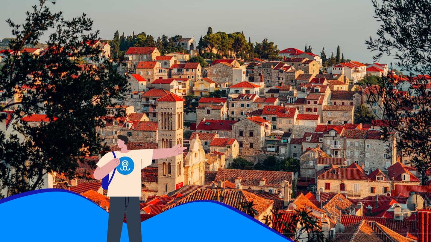 Pogled na bijele kuće s crvenim krovovima grada Hvara, uokviren lišćem drveća i grafikama Ferryscannera.