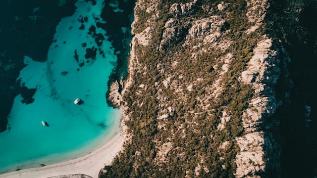 Veduta aerea della spiaggia di Roccapina in Corsica, con sabbia bianca, acqua turchese e promontorio roccioso coperto di macchia mediterranea.