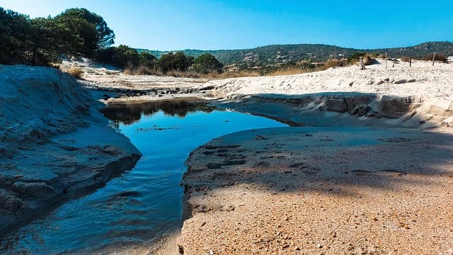 Sentiero sabbioso e piccolo corso d’acqua su una spiaggia della Corsica meridionale, circondata da colline verdi.