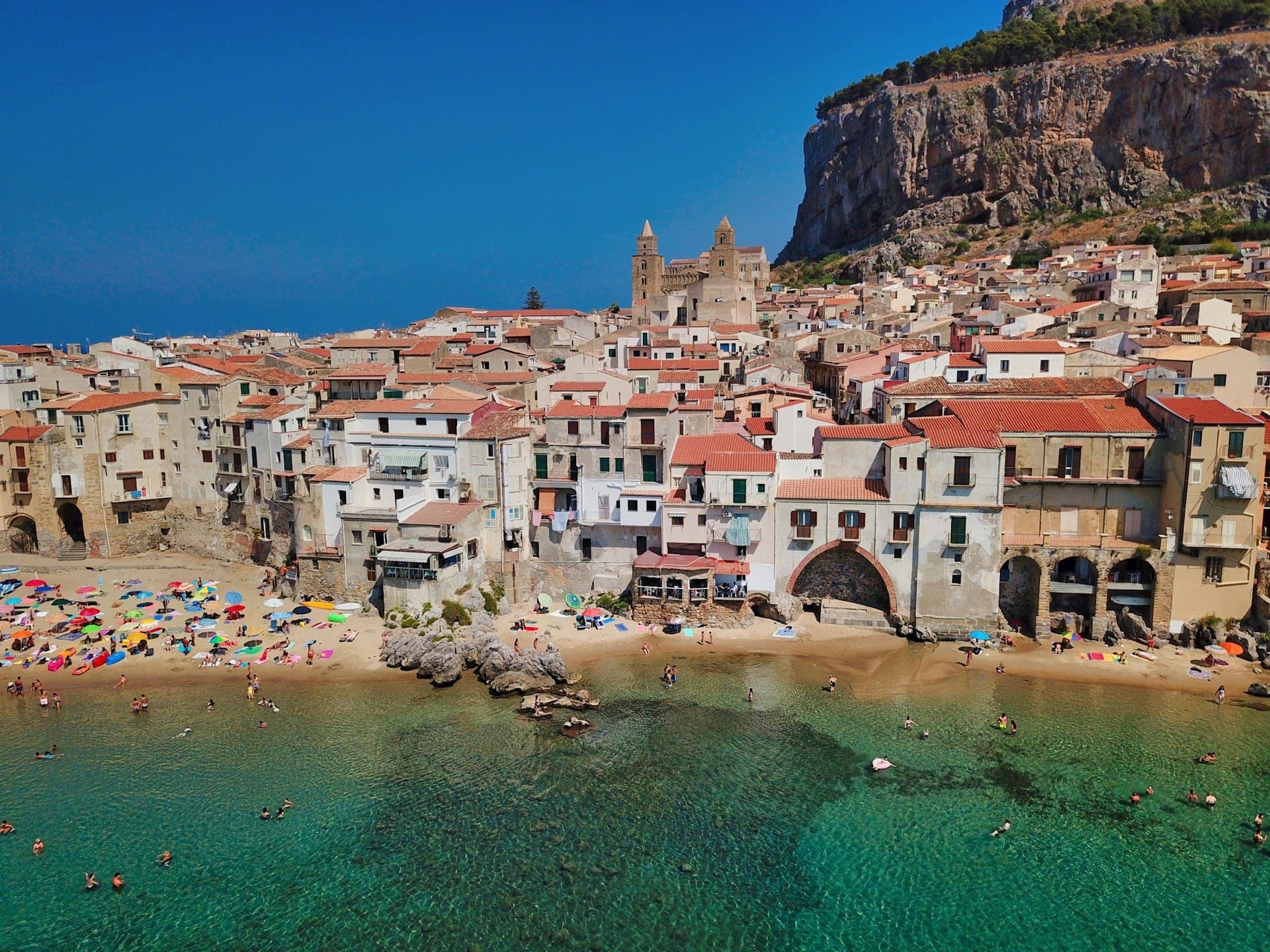 Petit village côtier devant une plage à l'eau transparente, avec une grande cathédrale et une falaise en toile de fond.