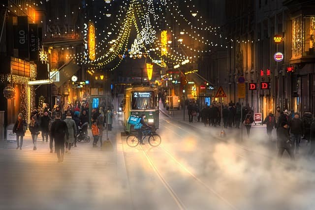Calle iluminada en el centro de la ciudad durante la Navidad en Helsinki, con tranvías, luces festivas y gente paseando entre tiendas y decoraciones.