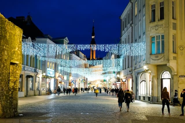 Strada pedonale nel centro di Tallinn illuminata da luci natalizie, con persone che passeggiano tra edifici storici al crepuscolo.