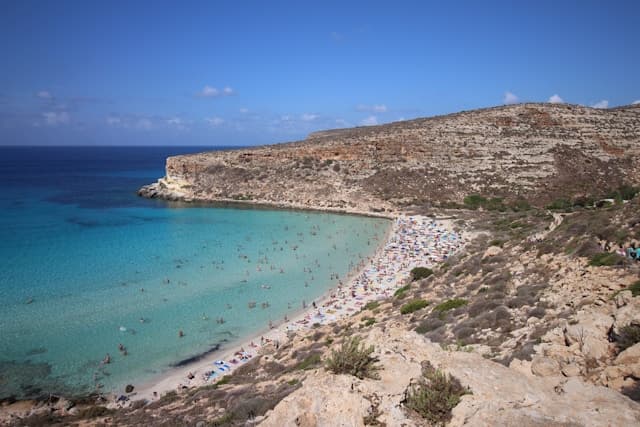 Spiaggia sabbiosa di Lampedusa con acqua turchese e fondali bassi, vista dall’alto con numerosi bagnanti lungo la costa.