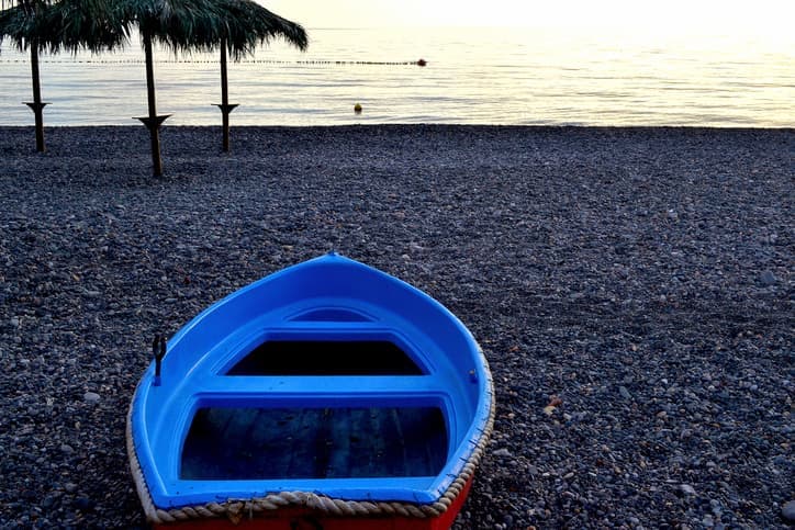 Barca azzurra sulla spiaggia di Marina Corta a Lipari, con ciottoli, mare calmo e ombrelloni in paglia sullo sfondo.