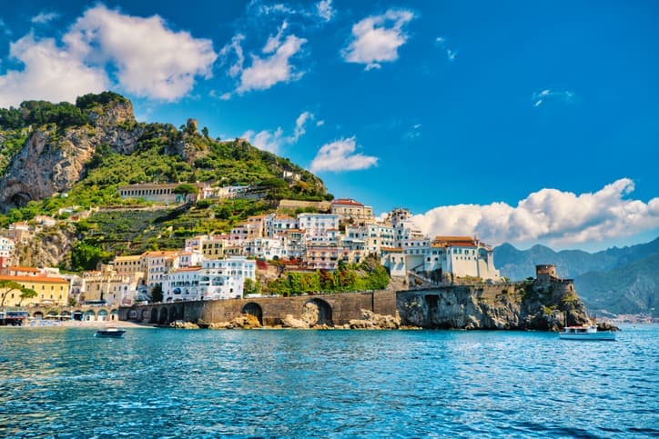 Vista dal mare di Amalfi, con le case bianche e colorate arrampicate sulla collina, l'arco in pietra sul porto e le montagne sullo sfondo.