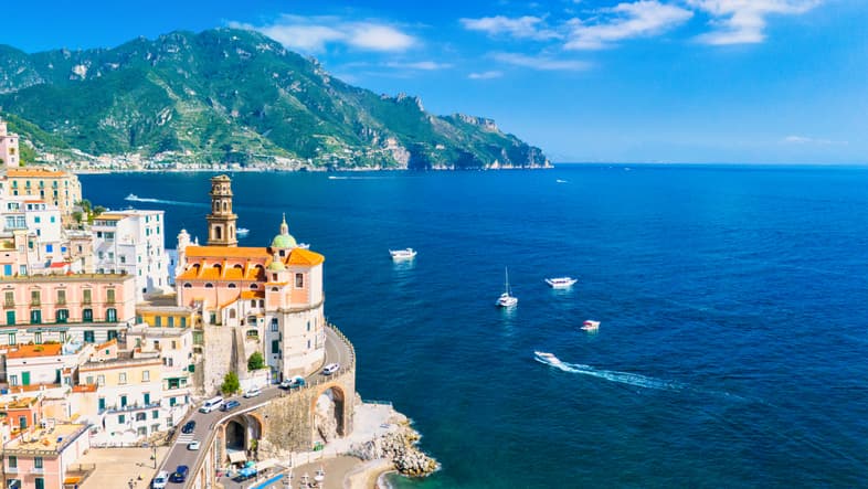 Vista aerea di Atrani sulla Costiera Amalfitana, con la Collegiata di Santa Maria Maddalena, le case colorate sulla scogliera e barche ancorate nel mare blu.