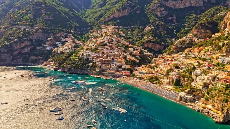 Vista aerea di Positano, con le case colorate arrampicate sulla collina, la Spiaggia Grande e il mare turchese pieno di barche.
