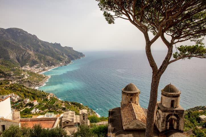 Vista panoramica da Ravello sulla Costiera Amalfitana, con un pino marittimo, le cupole di una chiesa e la costa che si estende fino al mare.